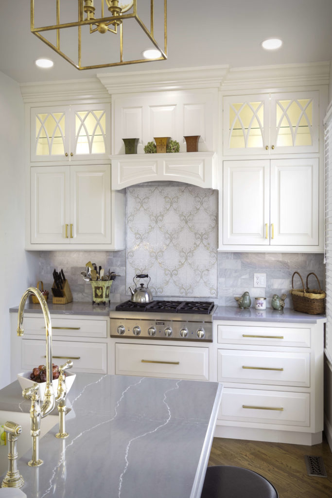 View of range area with glass upper cabinets and custom backsplash. Cabinets feature inset doors, under-cabinet power with dimmable lighting, and custom crown moulding.